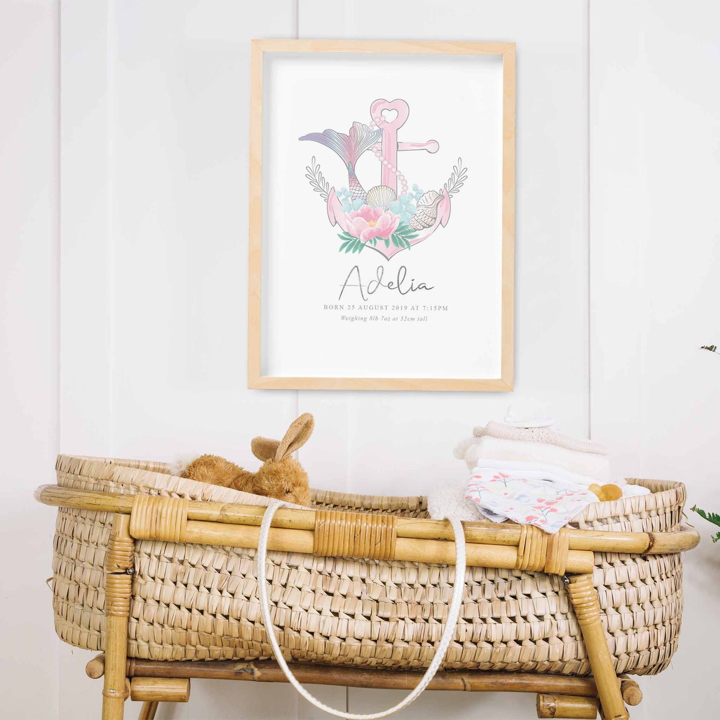 An oak framed picture of a pink nautical birth print featuring an anchor, mermaid tail, seashells, string of pearls and floral details decorated in a baby's nursery