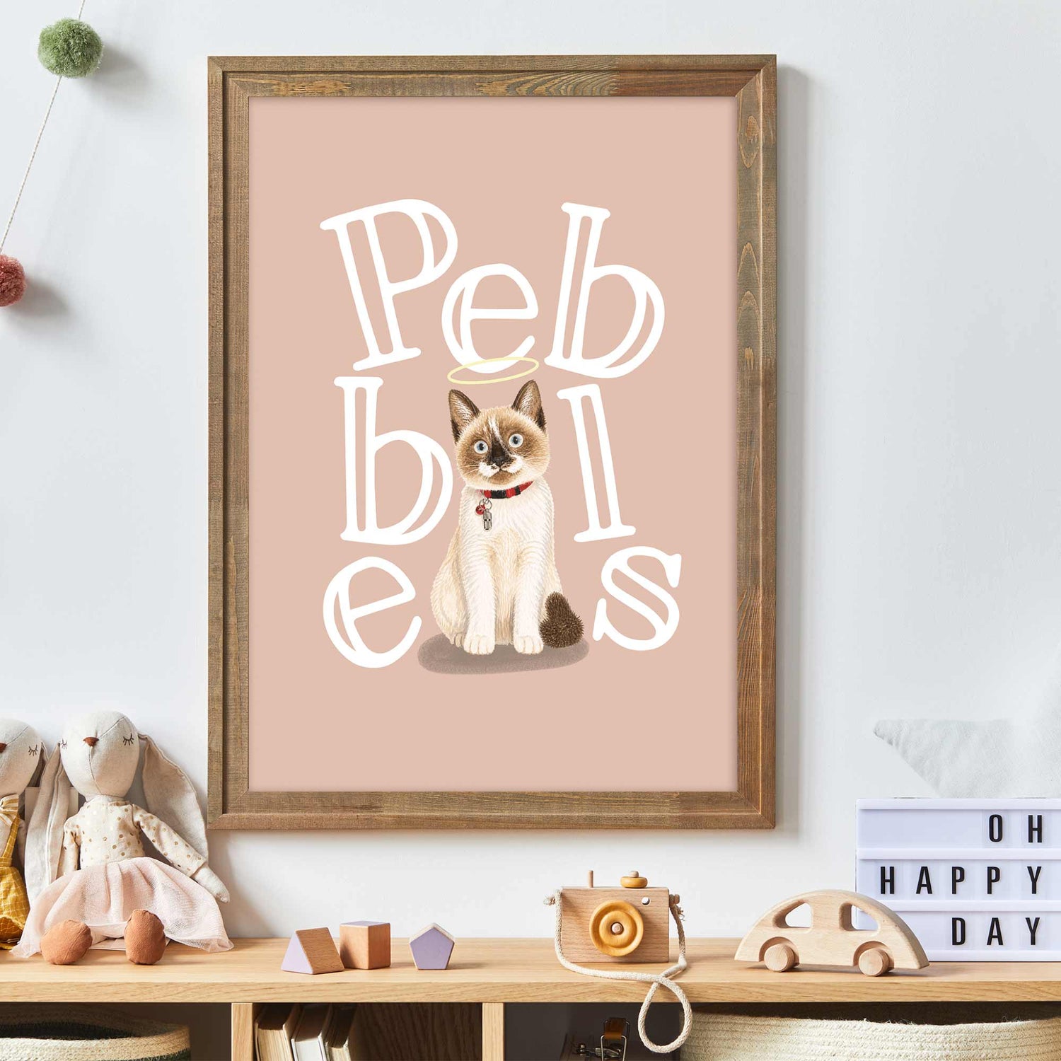 Framed kids pet portrait  with a cat illustration and 'Pebbles' text on a pink background, displayed on a shelf with toys.