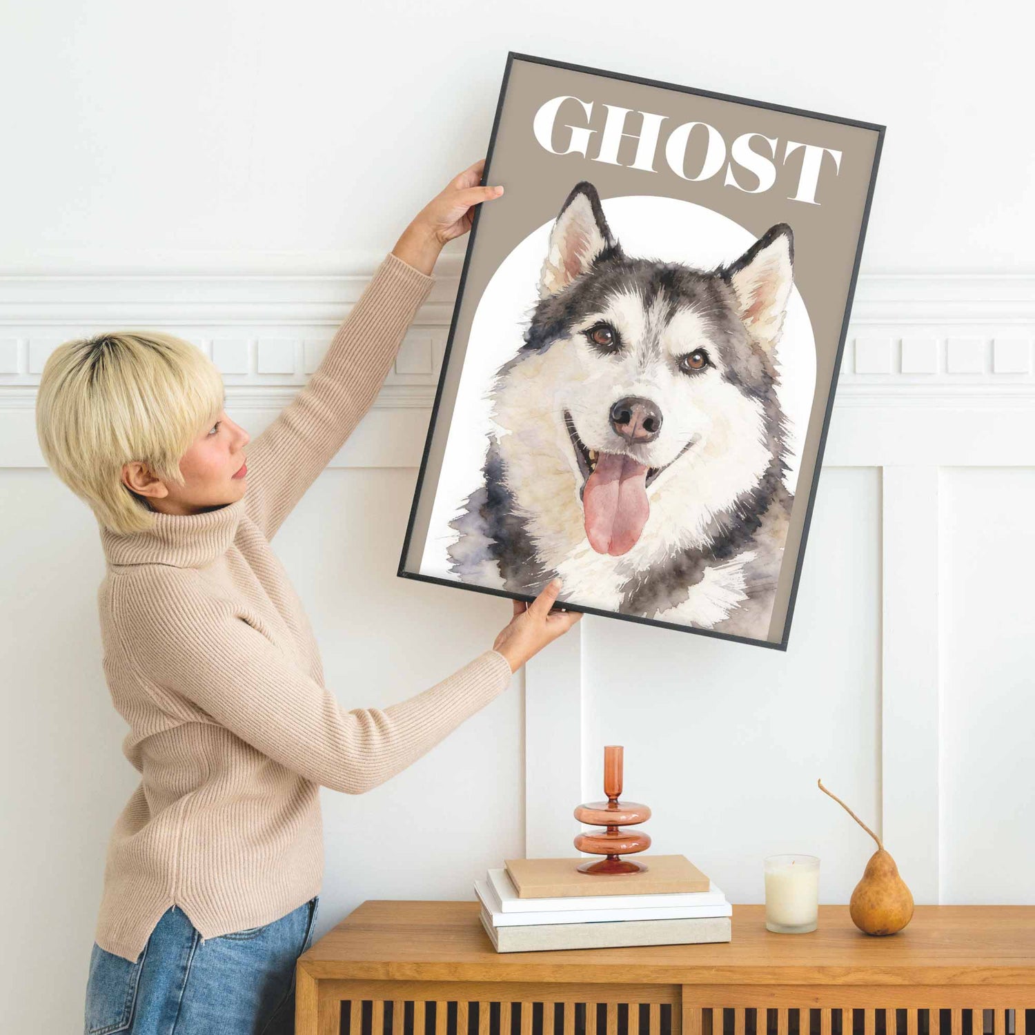 Person holding a framed picture of a dog portrait named 'Ghost' in a room with a wooden cabinet and decor items.