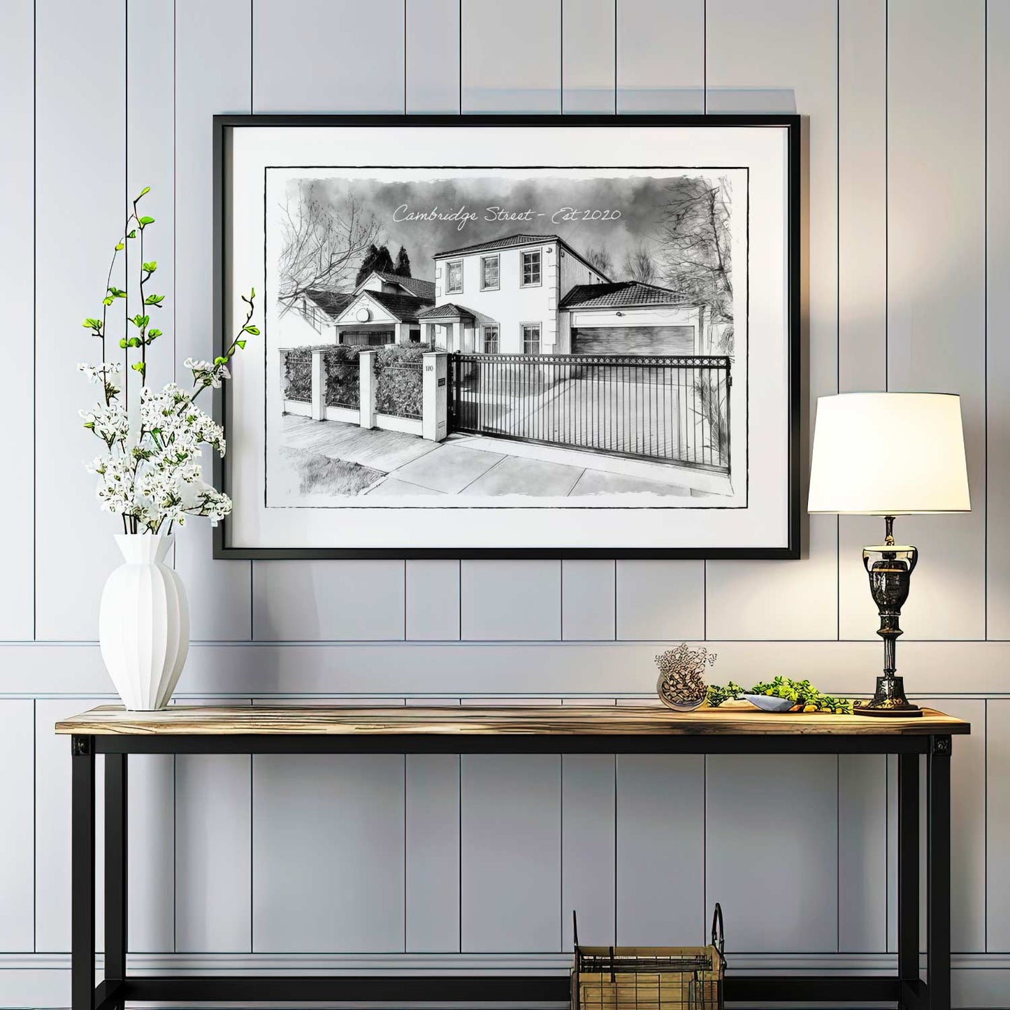Framed black and white custom home portrait artwork on a wall above a console table with decorative items.