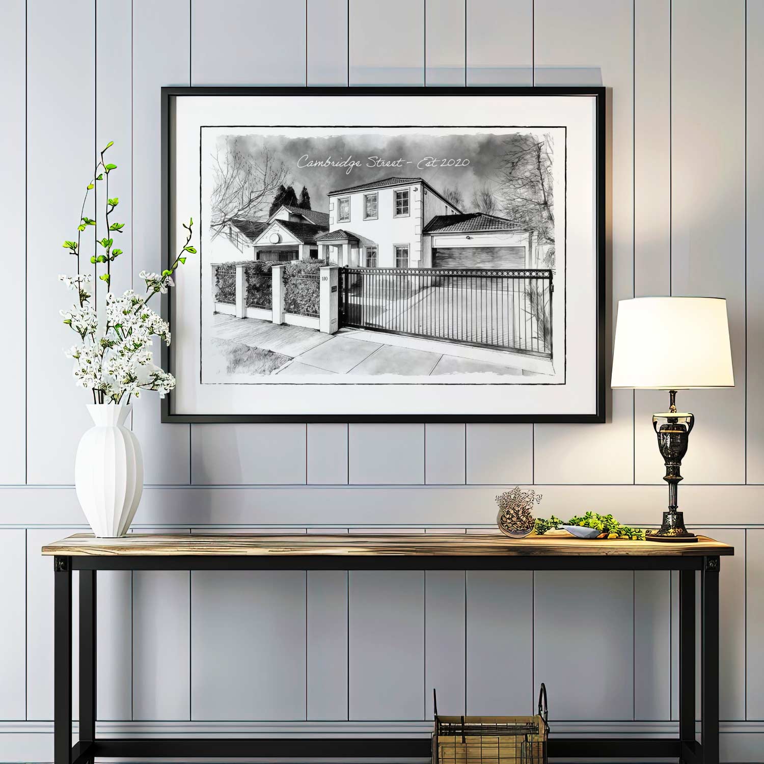 Framed black and white custom home portrait artwork on a wall above a console table with decorative items.