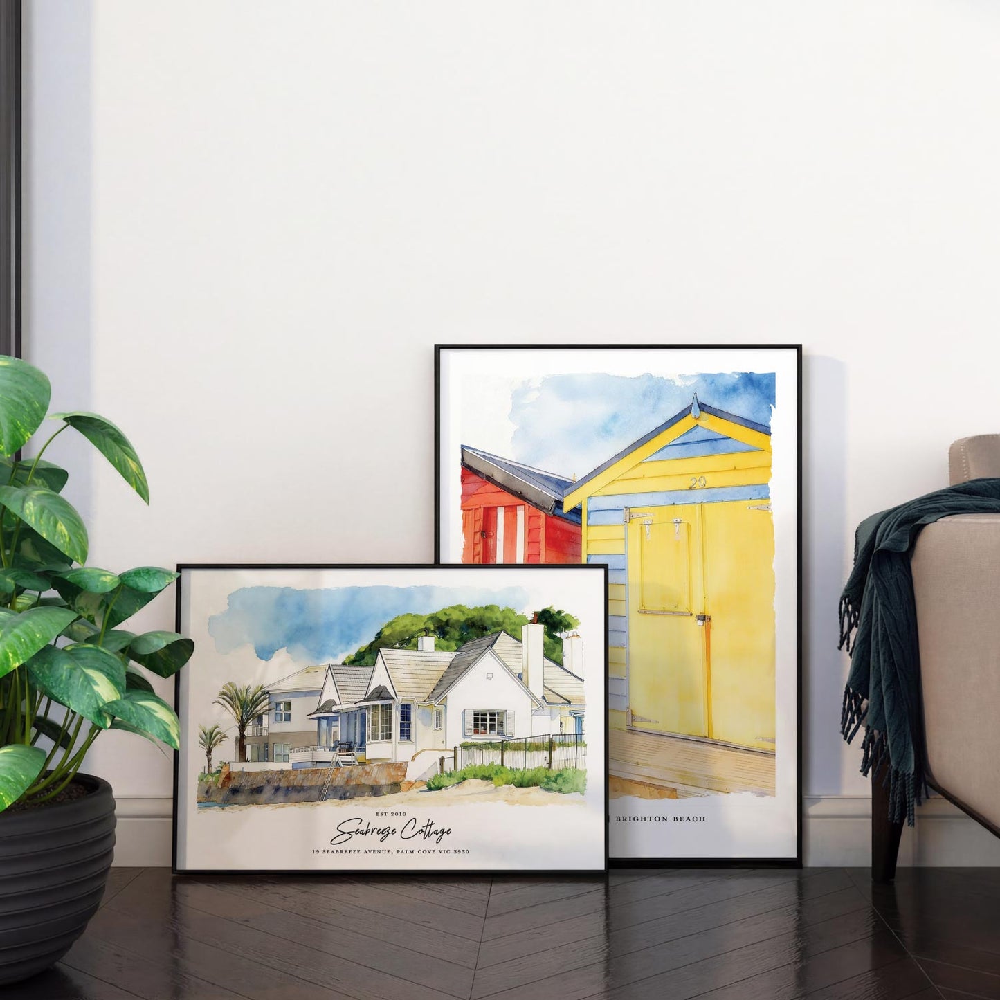 Two framed artworks of custom beach house portrait and custom beach box portrait on a wooden floor with a plant and chair in the background.