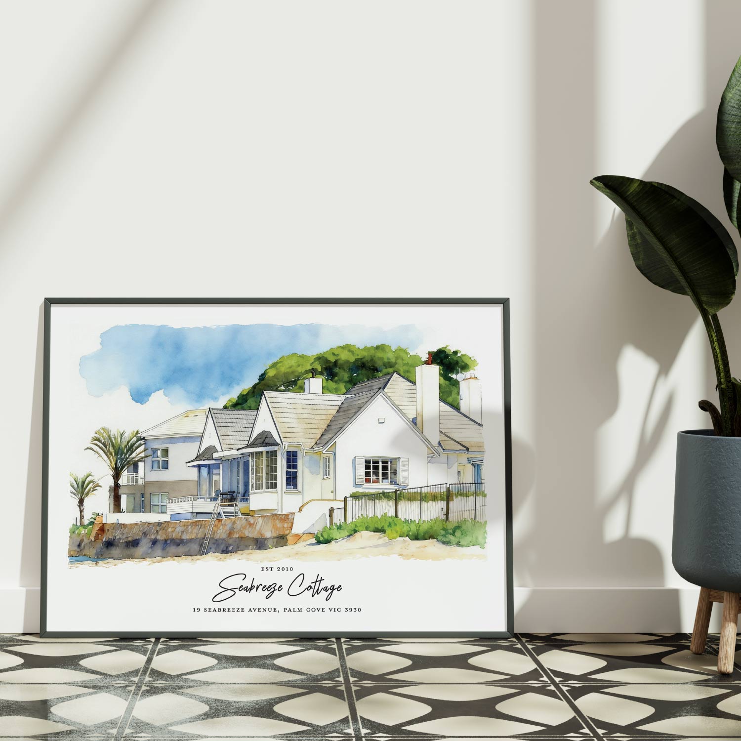 Framed artwork of a custom beach house portrait on a white wall with a plant to the right.