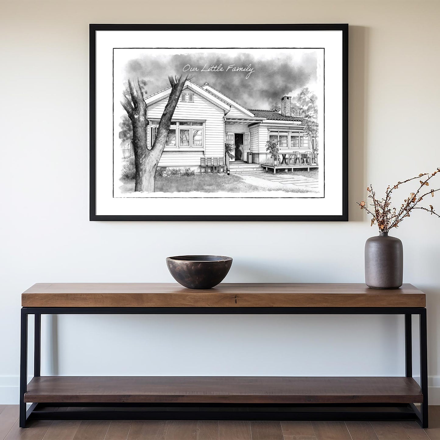 Framed black and white custom home portrait print on a wall above a wooden console table with decorative items.
