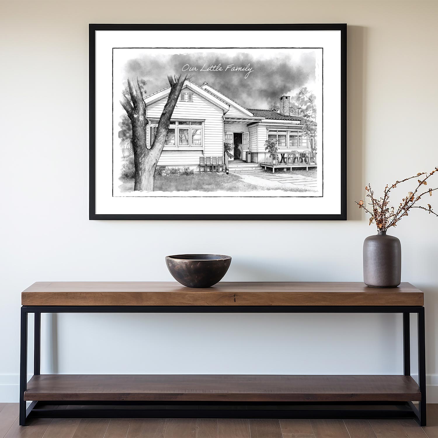 Framed black and white custom home portrait print on a wall above a wooden console table with decorative items.