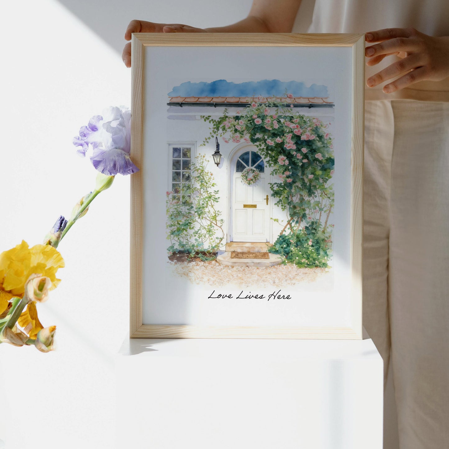 Framed artwork of a custom front door home portrait of a door with flowers and 'Love Lives Here' text, held by a person.