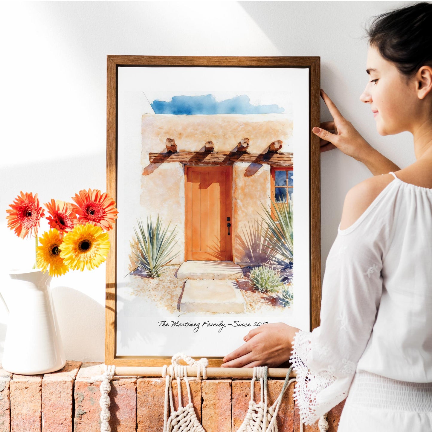 Woman holding a framed print of a custom front door home portrait of a door with decorative elements, next to a vase with flowers.