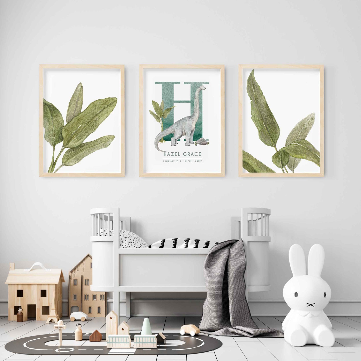 A set of 3 wall oak framed images of palm leaves and a dinosaur personalised monogram birth print displayed on the wall of a kids room