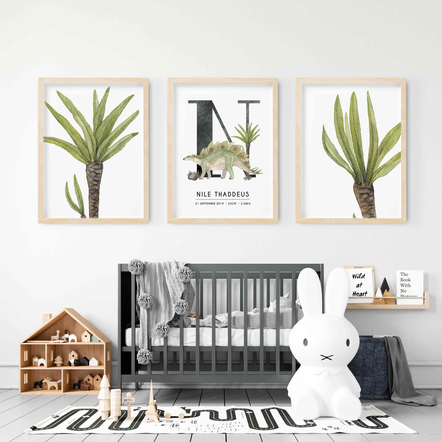 A set of 3 wall oak framed images of palm trees and a dinosaur personalised monogram birth print displayed on the wall of a kids room