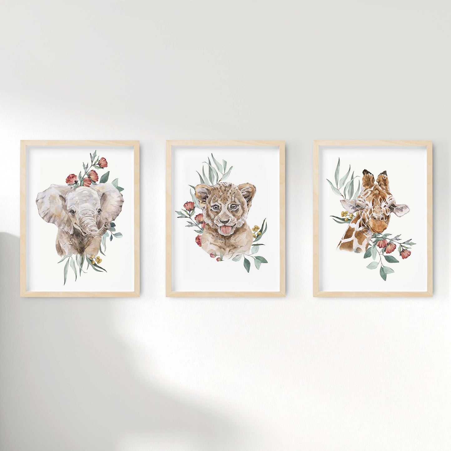 A set of 3 framed oak frames displaying kids nursery wall prints of a baby elephant, lion cub and baby giraffe on a botanical floral background